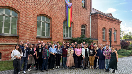 Sehr große Gruppe von Menschen steht vor dem Ministerium. Im Hintergrund eine Regenbogenfahne.