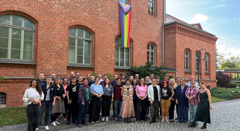 Sehr große Gruppe von Menschen steht vor dem Ministerium. Im Hintergrund eine Regenbogenfahne.